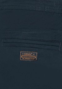 Un tissu bleu marine avec une texture lisse présente une poche arrière, des détails cousus et une étiquette en cuir marron sur laquelle est inscrit "SIGNAL".