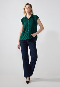 Grønn plissert kortermet bluse med knapper foran, sammen med mørkeblå rette jeans og svarte høye hæler.