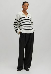 Gebreide trui in het wit met zwarte strepen, voorzien van een kraag en een relaxed fit, gecombineerd met zwarte wijd uitlopende broek en zwarte instappers.