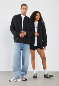 Black zip-up bomber jackets, loose blue jeans, and contrasting footwear. The model on the right wears black shorts and white socks.