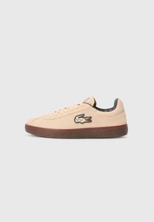 Baskets basses beiges avec semelle en caoutchouc foncé et logo crocodile vert sur le côté, posées sur un fond gris clair uni.