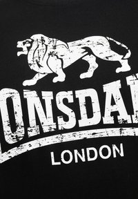 Sort bomulds t-shirt med et slidt hvidt grafik af en løve og teksten "Lonsdale London" i fed, stiliseret skrifttype.