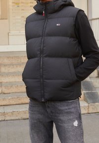 Svart puffer-väst med hög krage, framdragkedja och logotyp. Bärs över en svart långärmad tröja och matchas med slitna grå jeans.