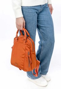 Orange nylon backpack with dual top handles, side pockets, zipper accents, and adjustable straps. Worn with light blue jeans and white sneakers.