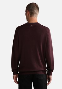 Maroone langærmet sweater i blød tekstur, med rund halsudskæring og lille logo-detalje i ryggen, parret med sorte bukser.