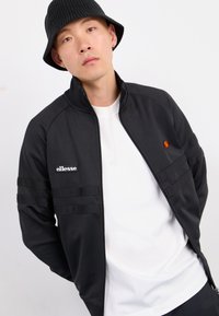 Młody mężczyzna w czapce typu bucket hat, czarnej kurtce na zamek z logo ellesse oraz białej koszulce, pozujący na białym tle.
