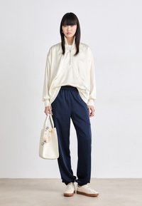 Off-white zip-up pūkinys su priekinėmis kišenėmis, derinamas su tamsiai mėlynomis elastinėmis kelnėmis. Priedai apima kremą spalvos rankinę ir smėlio spalvos sportbačius.
