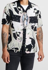 Černá košile s krátkým rukávem s květinovými a křížovými vzory, která má uprostřed ilustraci postavy. Vyrobena z hladkého, lehkého materiálu.