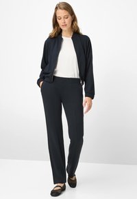 Marineblaue Bomberjacke, weißes Oberteil und marineblaue Hose kombiniert mit schwarzen Mesh-Ballerinas. Glatter Stoff, lockere Passform, elastische Bündchen und gerade geschnittene Beine.