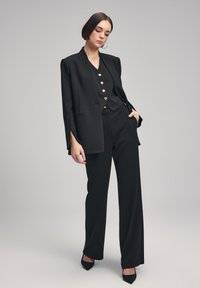 Blazer negro ajustado con hombros estructurados, combinado con una blusa negra de botones y pantalones de pierna ancha a juego. Acabado con botones dorados.