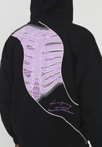 Sweat à capuche noir avec un grand motif de cage thoracique violette sur le dos, avec un contour courbé et des détails blancs, fabriqué en coton doux.