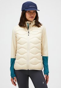 Gilet imbottito beige con motivo a cuciture ondulate, chiusura con zip e tasche laterali, indossato sopra una maglietta chiara con maniche teal. Un berretto scuro completa il look.