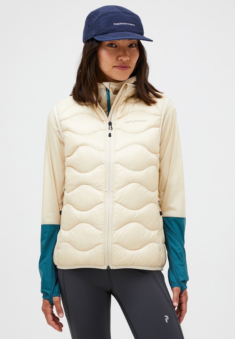 Beige puffer vest met golvende quiltpatroon, rits aan de voorkant en zijzakken, gedragen over een lichte top met teal mouwen. Donkere pet maakt de look af.