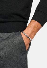Silberarmband mit Kettengliedern an einem Handgelenk, bestehend aus ineinandergreifenden Gliedern und einem Verschluss, getragen zu grauer Stoffhose und schwarzem Pullover.
