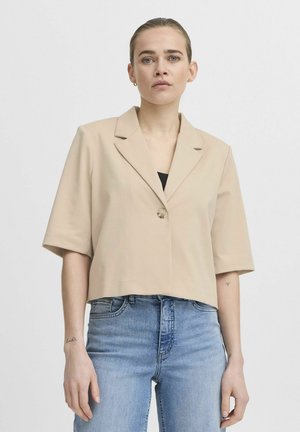 Beige cropped blazer dengan lengan pendek, penutupan kancing tunggal, dan kerah notch, dipadukan dengan celana jeans denim biru. Tekstur kain yang halus.