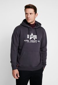 Sudadera gris de mezcla de algodón, con un bolsillo frontal y diseño de logo blanco. La tela parece suave con una textura lisa.