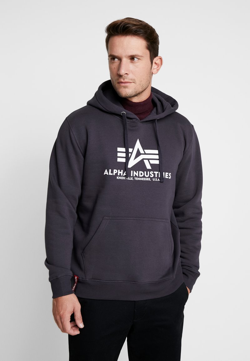 Sudadera gris de mezcla de algodón, con un bolsillo frontal y diseño de logo blanco. La tela parece suave con una textura lisa.