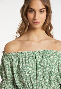Top verde floreale con scollatura a spalle scoperte e colletto arricciato, realizzato in un leggero tessuto testurizzato, decorato con piccoli fiori bianchi su uno sfondo verde.