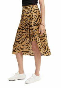 Vrouw in een beige en zwarte dierenprint asymmetrische midi-jurk, gecombineerd met witte sneakers en een zwart top.