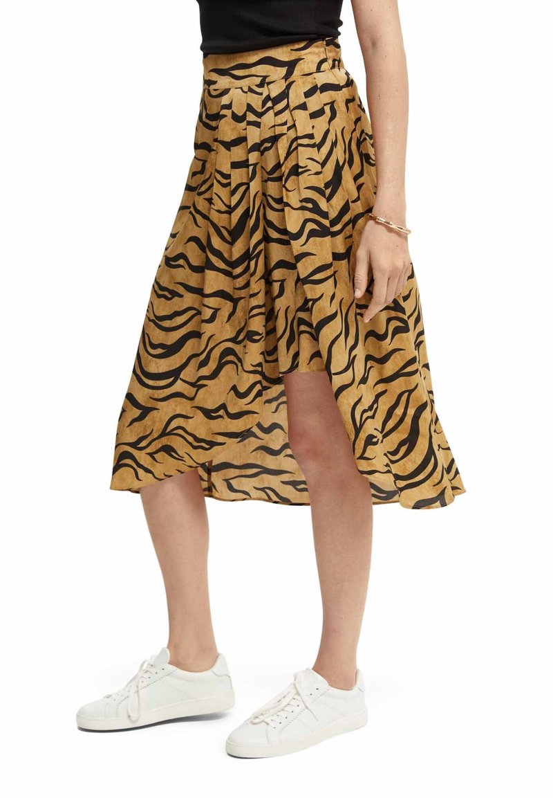Vrouw in een beige en zwarte dierenprint asymmetrische midi-jurk, gecombineerd met witte sneakers en een zwart top.