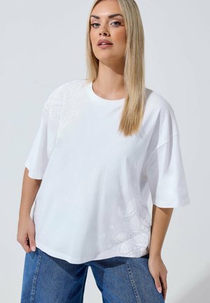 Femme aux longs cheveux blonds portant un t-shirt blanc ample avec des motifs floraux subtils et un jean bleu, debout devant un fond uni.