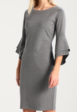 Kobieta w szarej obcisłej sukience do kolan z warstwowymi rękawami dzwonkowymi i szerokim, zaokrąglonym dekoltem, stojąca na jednolitym tle.