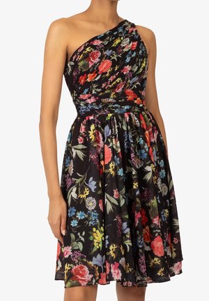 Ein schwarzes One-Shoulder-Kleid mit farbenfrohem Blumenmuster, plissiertem Oberteil und geraffter Taille, getragen von einer stehenden Person.