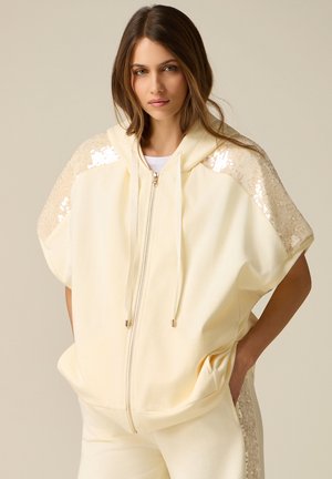 Donna che indossa una giacca con cappuccio crema con zip e dettagli di paillettes sulle spalle e pantaloni abbinati, mani in tasca, sfondo neutro.