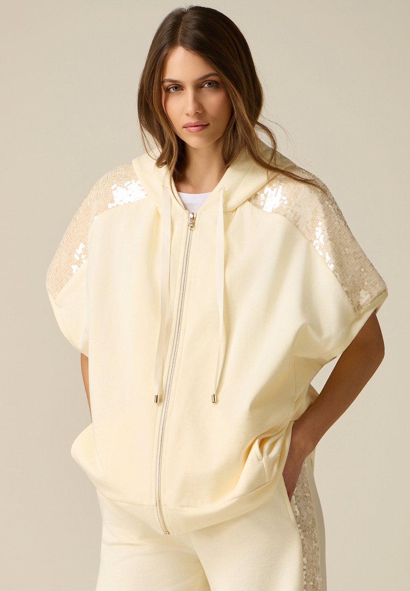 Mujer con chaqueta con capucha cremosa con cremallera y detalles de lentejuelas en los hombros y pantalones a juego, manos en los bolsillos, fondo neutro.