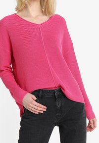 Różowy sweter z dzianiny z dekoltem w serek i oversize'owym krojem, z opuszczonymi ramionami i długimi rękawami, zestawiony z czarnymi dżinsami.