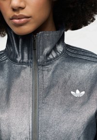 Traper jakna s visokim ovratnikom, metalik tamnoplave boje, zatvaranje na patentni zatvarač i bijeli izvezeni logo na lijevoj strani prsa. Teksturirani materijal.