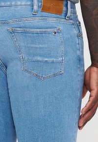 Jeans in denim blu chiaro con una texture spazzolata, dotati di una tasca posteriore e di una patch in pelle marrone con logo in vita.