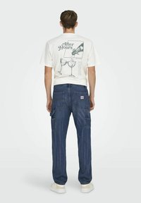 T-shirt blanc avec impression graphique au dos ; pantalon cargo en denim bleu avec plusieurs poches ; modèle portant des baskets blanches.