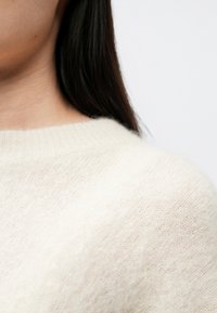 Pehme ja kreemjat värvi kampsun soonikulise kaelusega, millel on tekstuuriga kootud muster ja veidi lõdvem lõige.