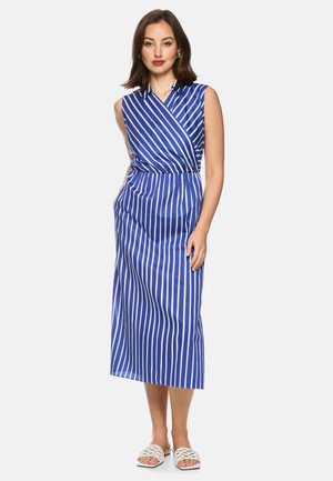 Woman standing wearing a sleeveless blue and white vertical striped midi dress and white woven slip-on sandals on a white background.