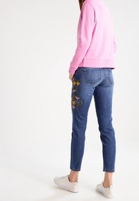Jeans ajustados de color azul con bordado floral en la pierna izquierda, hechos de tela de mezclilla. Combinados con una sudadera rosa y zapatillas blancas.
