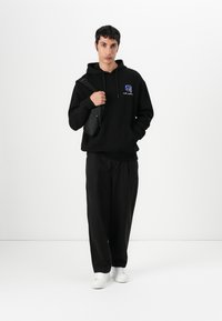Felpa nera con grafica ricamata, abbinata a pantaloni neri larghi e sneakers bianche. Il modello tiene una piccola borsa a tracolla.