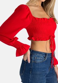 Blusa roja corta con textura fruncida, mangas abullonadas anchas y dobladillo con volantes. Combinada con jeans de mezclilla azul de talle alto.