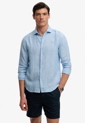 Jasnoniebieska koszula w paski na guziki z długimi rękawami, zestawiona z ciemnymi szortami. Posiada małe logo na piersi oraz usztywniany kołnierzyk.