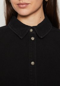 Camicia in denim nero con colletto a punta, cinque bottoni argentati e cuciture visibili. Il tessuto ha una texture liscia.