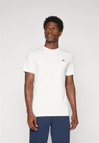 Λευκό βαμβακερό T-shirt με κοντά μανίκια, στρογγυλή λαιμόκοψη και ένα μικρό πράσινο λογότυπο στο αριστερό στήθος. Φορεμένο με ναυτικές παντελόνες.