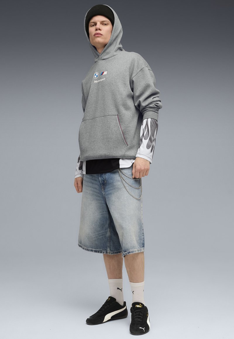 Grå hætte sweatshirt med logo, lagdelt over en sort t-shirt, parret med blå denim shorts. Sorte sneakers og hvide sokker med logo accenter.