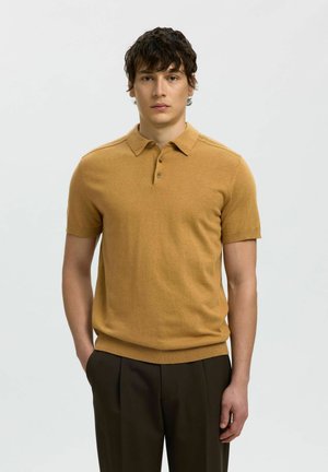 Jeune homme aux cheveux courts et foncés, portant un polo à manches courtes couleur moutarde et un pantalon marron foncé, debout avec une main dans la poche.