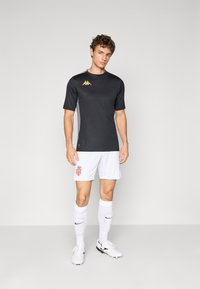 T-shirt de sport noir à manches courtes avec un logo doré, associé à un short blanc arborant un emblème rouge et doré. Chaussettes blanches et chaussure noire.