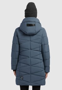 Padded coat v sivom modrom odtieni, s geometrickým prešívaným dizajnom, kapucňou a čiernym logom na spodnom leme. Na vrchu je textúrovaná pletená čiapka.