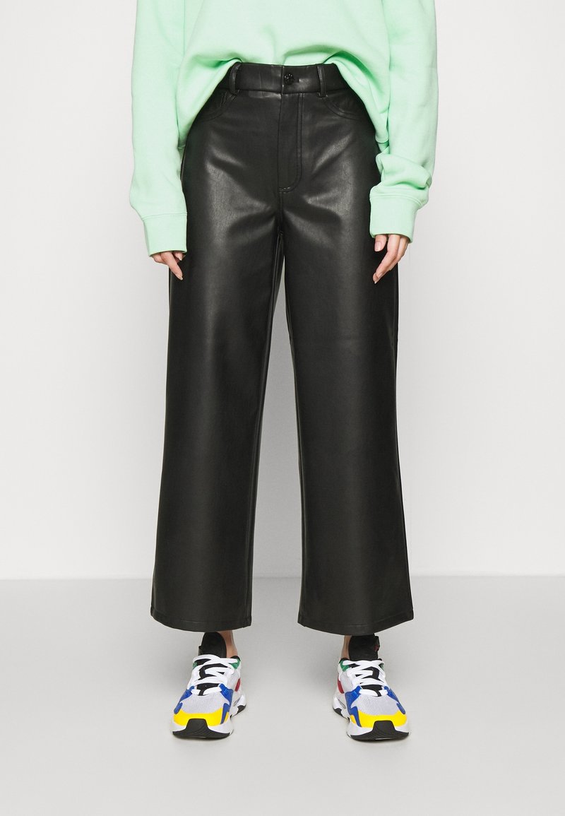 Personne portant un pantalon en cuir noir à jambes larges, une chemise manches longues vert clair et des baskets blanches colorées, debout sur un fond uni.