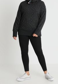 Persona che indossa un hoodie nero a pois, jeans skinny neri e sneakers bianche con piattaforma, in piedi contro uno sfondo chiaro e uniforme.