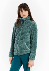 Mlada žena s dugom smešnom kosom, obučena u teletu kaput sa patent zatvaračem i teletu hlače, stoji ispred obične bele pozadine.