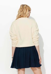 Femme blonde de dos portant un pull crème à manches longues et une jupe plissée bleu foncé sur fond blanc uni.