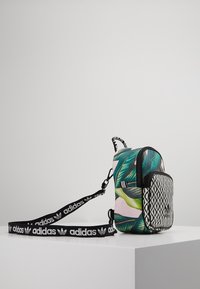 Wielokolorowy plecak z tropikalnym wzorem liści i czarną geometryczną kieszenią z przodu. Zawiera mocny czarny pasek z detalami logo.